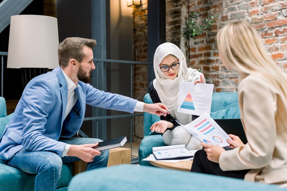The Role of Branding Agencies in Building a Memorable Brand Identity three multiracial male and female business people sitting on couch in the modern office room with decorative brick wall and discussing the development and main results of big business project